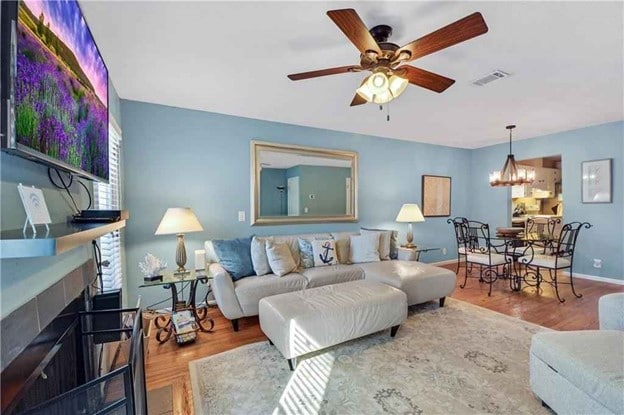 A living area features a light-colored sectional sofa facing a mounted television, with a large mirror on the wall enhancing the sense of space. Two lamps provide soft lighting, while a dining area with an iron table and four chairs is visible in the background.