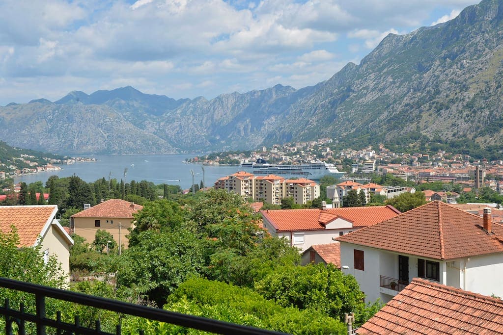 A scenic view of Kotor Bay is captured from the balcony, showcasing the tranquil waters bordered by lush greenery and mountains. The charming rooftops of nearby houses add a touch of local architecture, while the distant town reveals a backdrop of stunning natural beauty.