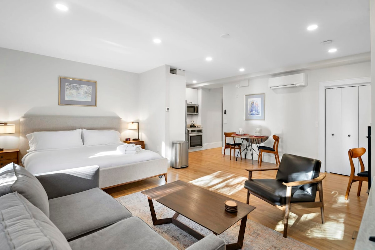An open studio layout is features a king bed and a queen sleeper sofa, complemented by a coffee table and two leather side chairs. A dining area is furnished with a drop leaf table and chairs. The modern kitchen includes stainless steel appliances, with natural light illuminating the space.