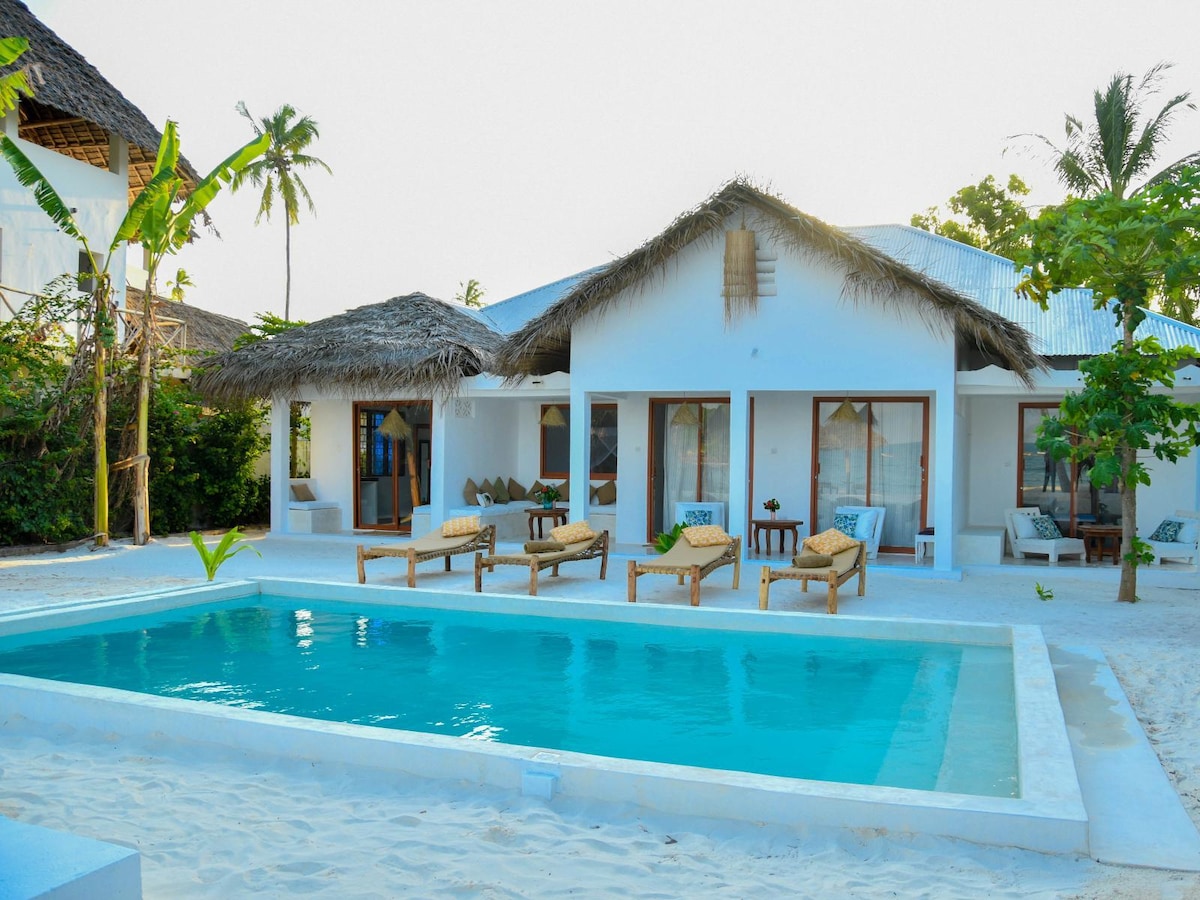 A beachfront property with a thatched roof features multiple sun loungers arranged around a serene, clear pool. Large glass doors provide views into the inviting indoor space. Lush tropical foliage and palm trees enhance the peaceful atmosphere of the outdoor area.