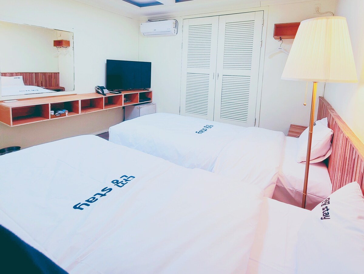 A well-lit room features two spacious beds covered with white linens, promoting a clean and comfortable atmosphere. A wooden dresser with a television and lamp is positioned across from the beds. Large windows allow natural light to brighten the space.