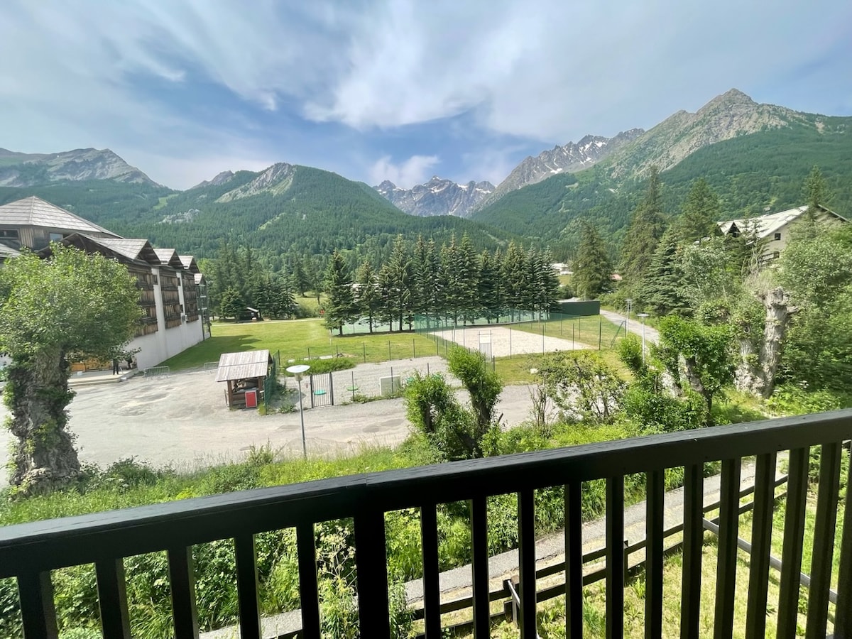 The balcony view captures a picturesque landscape of mountains and greenery. A spacious field featuring volleyball courts is visible, with tennis courts in the background. The natural surroundings offer a serene atmosphere, enhancing the outdoor experience from the apartment.
