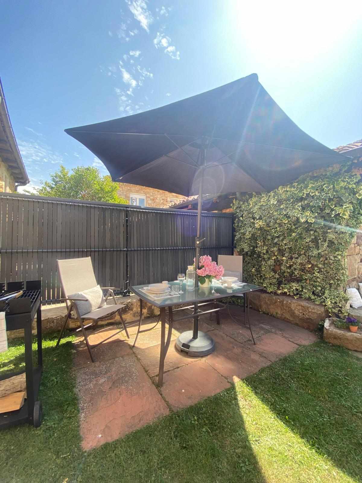 An outdoor garden area features a table set for four, shaded by a large parasol, surrounded by greenery and a wooden fence. Two chairs are positioned at the table, and a portable barbecue is located nearby on a paved surface.