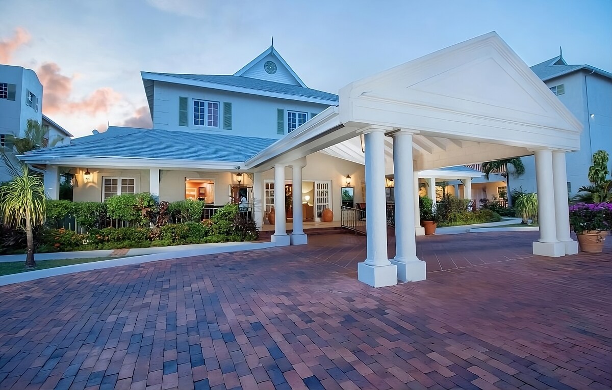 The entrance to the resort features a covered portico supported by white columns, leading to the welcoming lobby. Lush landscaping with tropical plants surrounds the building, accented by soft evening light and a blue sky.