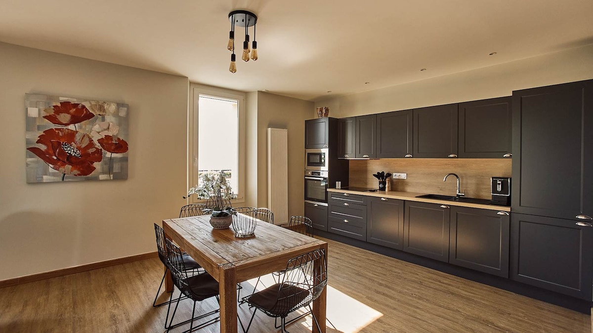 A modern kitchen area features dark cabinetry and a light wood countertop. The space includes a dining table with black wire chairs, complemented by a wall art piece depicting red flowers. Large windows provide natural light, enhancing the inviting atmosphere.