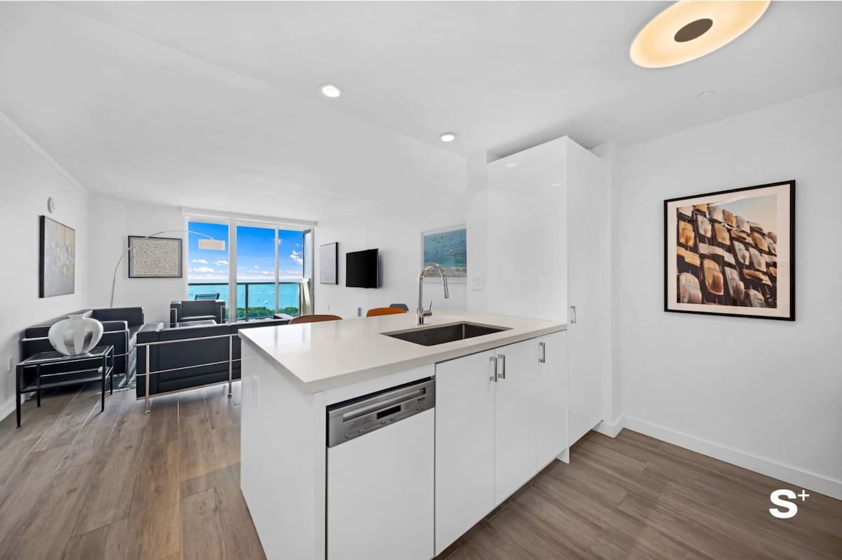 A modern kitchen area with sleek white cabinetry and a light-colored countertop is shown. The open layout leads to a bright living room featuring large windows that offer views of the outdoors. Minimalist artwork adorns the walls, contributing to a clean aesthetic.