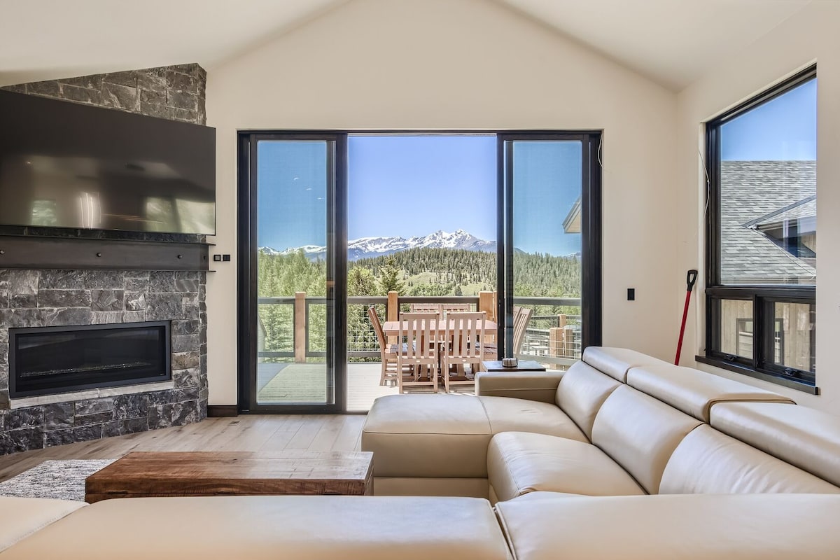 A modern living area is highlighted by a large sectional sofa facing a sleek stone fireplace. Expansive glass doors provide unobstructed views of the surrounding mountains and outdoor dining space, enhancing the connection between indoor and outdoor settings.
