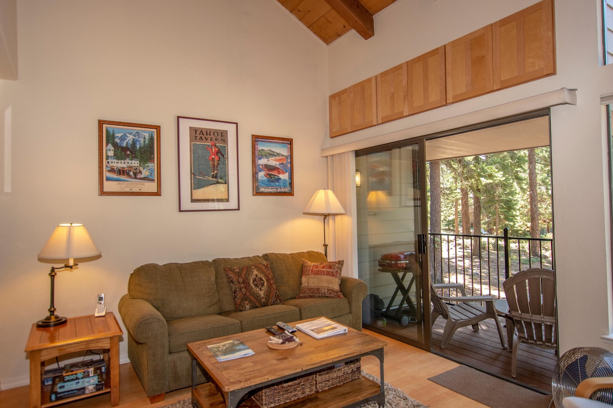 A spacious living area features a comfortable sofa, accompanied by a coffee table and side lamp. Walkout access is provided to a deck with outdoor seating and views of surrounding trees. Wall art depicting local scenes enhances the room's character.