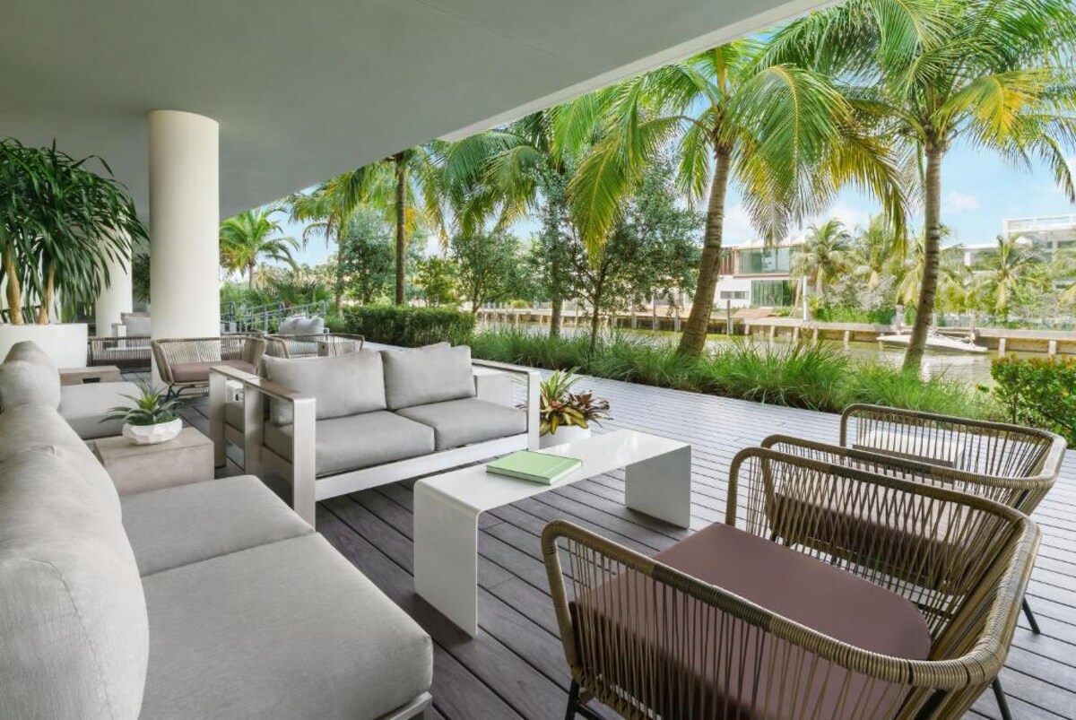 A spacious outdoor seating area is surrounded by tropical plants and palm trees. Comfortable grey sofas and woven chairs are arranged on a wooden deck, inviting relaxation. Natural light enhances the tranquil atmosphere, creating an ideal space for enjoying the views of the neighborhood.
