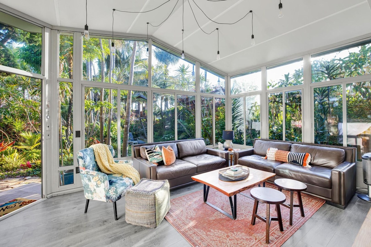 The living room features floor-to-ceiling windows that provide a view of the lush, tropical landscape outside. Two comfortable sofas and a patterned area rug are arranged around a coffee table. A colorful accent chair and pouf add a touch of style to the space.