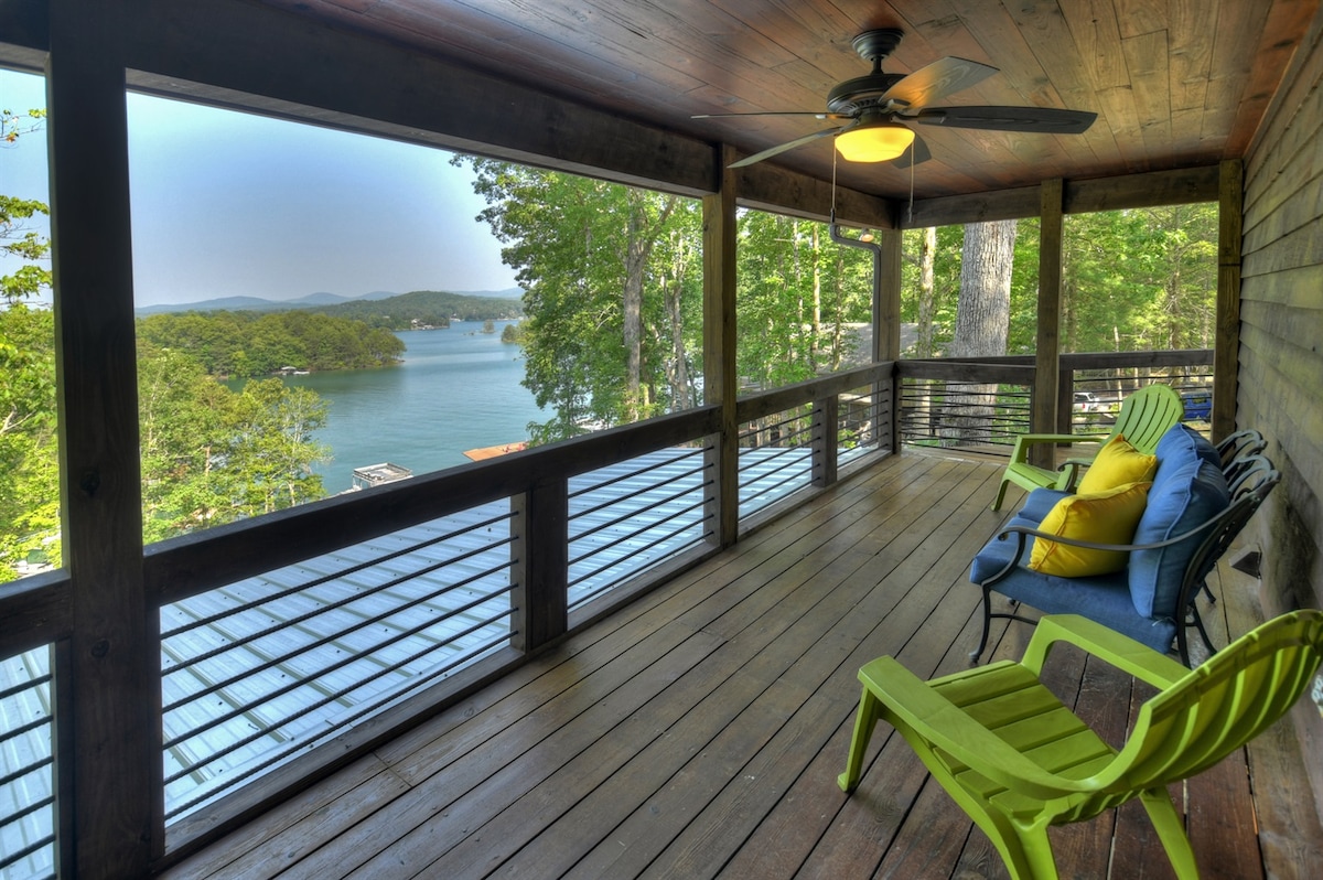 A covered porch features comfortable seating with bright green and blue chairs, offering views of the lake and surrounding trees. A ceiling fan hangs above, enhancing the outdoor space. The wooden flooring and railing create a natural, inviting ambiance.