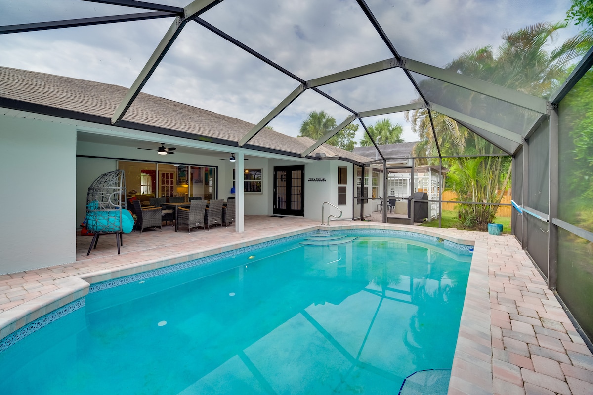 A screened-in swimming pool area features clear water reflecting the sky above. Surrounding the pool, paver stones create an inviting space, with lounge seating visible under the shaded lanai. The area is surrounded by lush greenery, enhancing the outdoor relaxation setting.