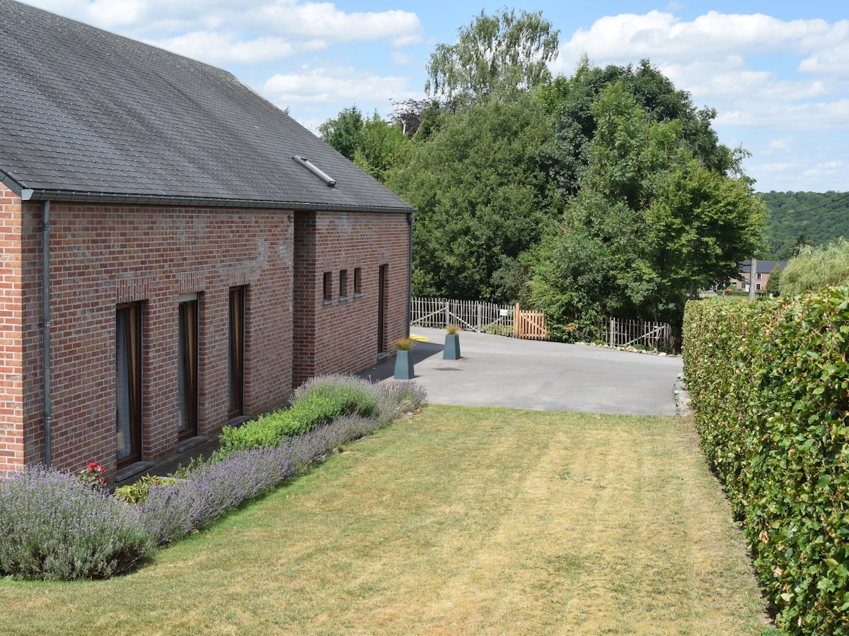 A contemporary building is set on a grassy lawn, surrounded by well-maintained shrubbery and flowering plants. The landscape features a stone pathway leading to a parking area, with a view of rolling greenery in the background under a clear sky.