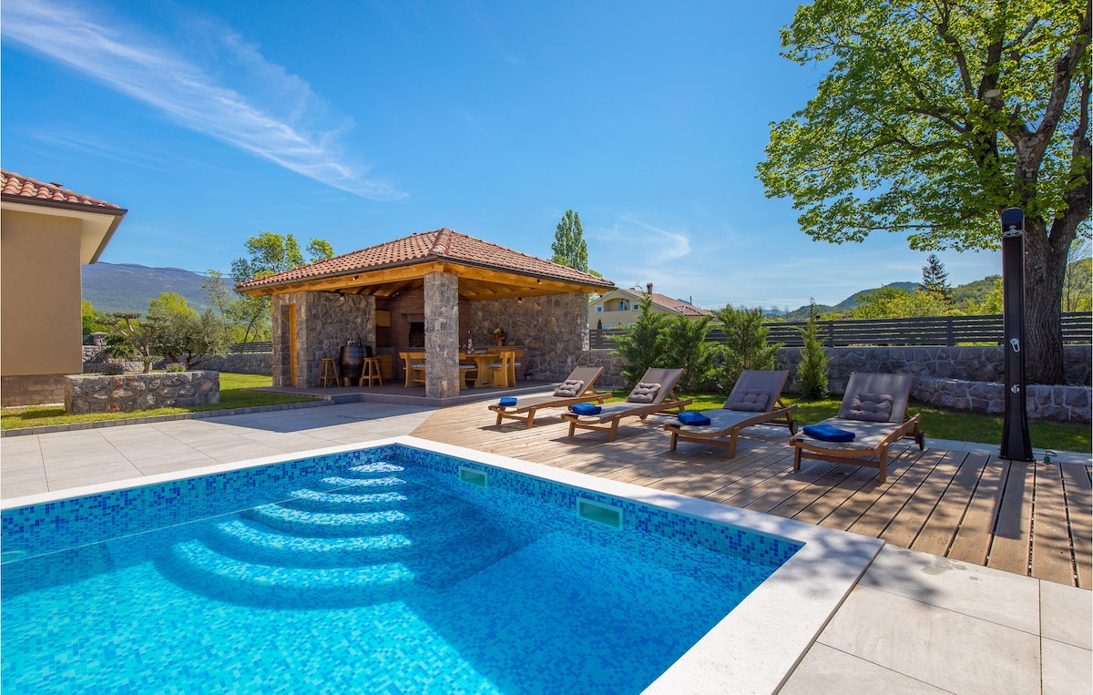 A private outdoor pool is set within a landscaped garden, accompanied by sun loungers arranged on a wooden deck. In the background, a covered outdoor kitchen and dining area are visible, surrounded by greenery and under a clear blue sky.