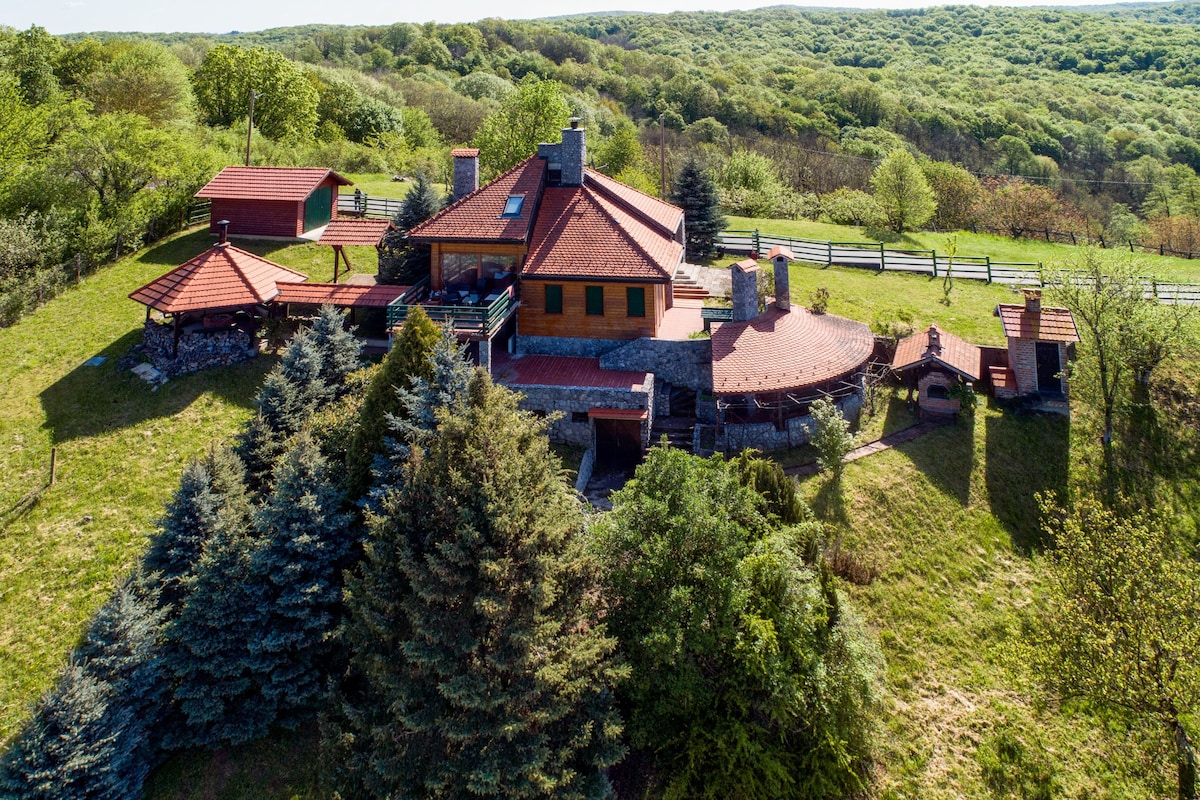 An aerial view of a rustic hunting lodge surrounded by lush greenery. The two-level structure features a mixture of red-roofed sections and natural stone, with open terraces and wooden accents. The landscape is dotted with trees and rolling hills, emphasizing a tranquil natural setting.