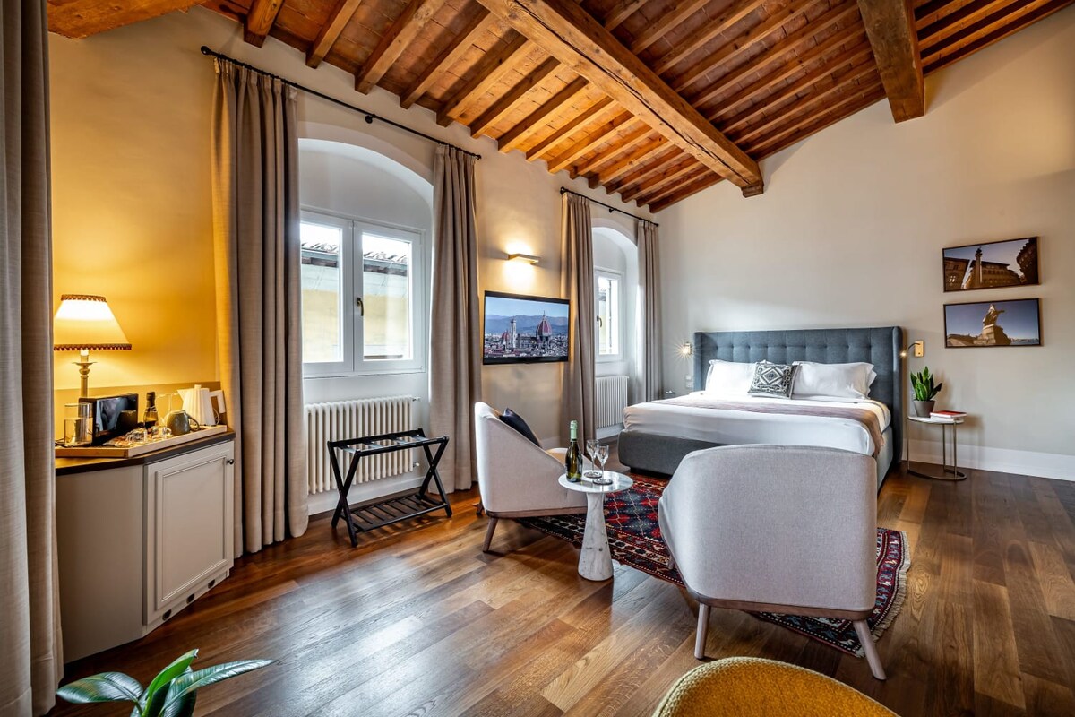 A spacious suite features wooden ceilings and soft lighting. A large bed is positioned centrally, with modern seating and a table in front. Nearby, a minibar is visible, complemented by natural light from arched windows. Artwork hangs on the walls, adding a touch of elegance.