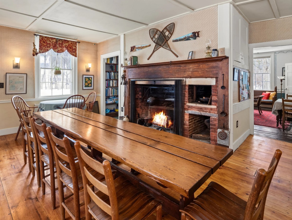 A long wooden table is surrounded by several wooden chairs in a warm dining area. A brick fireplace is centrally located, with a fire glowing inside. The walls display bookshelves and decorative items, enhancing a communal gathering space.