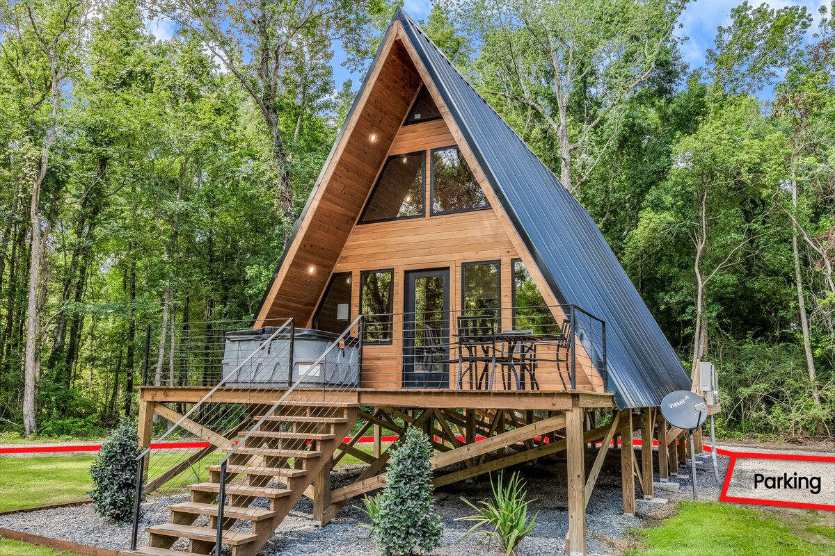 A modern A-Frame cabin is nestled among lush greenery, featuring a sleek metal roof and expansive windows. A spacious deck is outlined with seating and a hot tub, offering a welcoming space to relax. Steps lead up to the entry, surrounded by manicured landscaping.