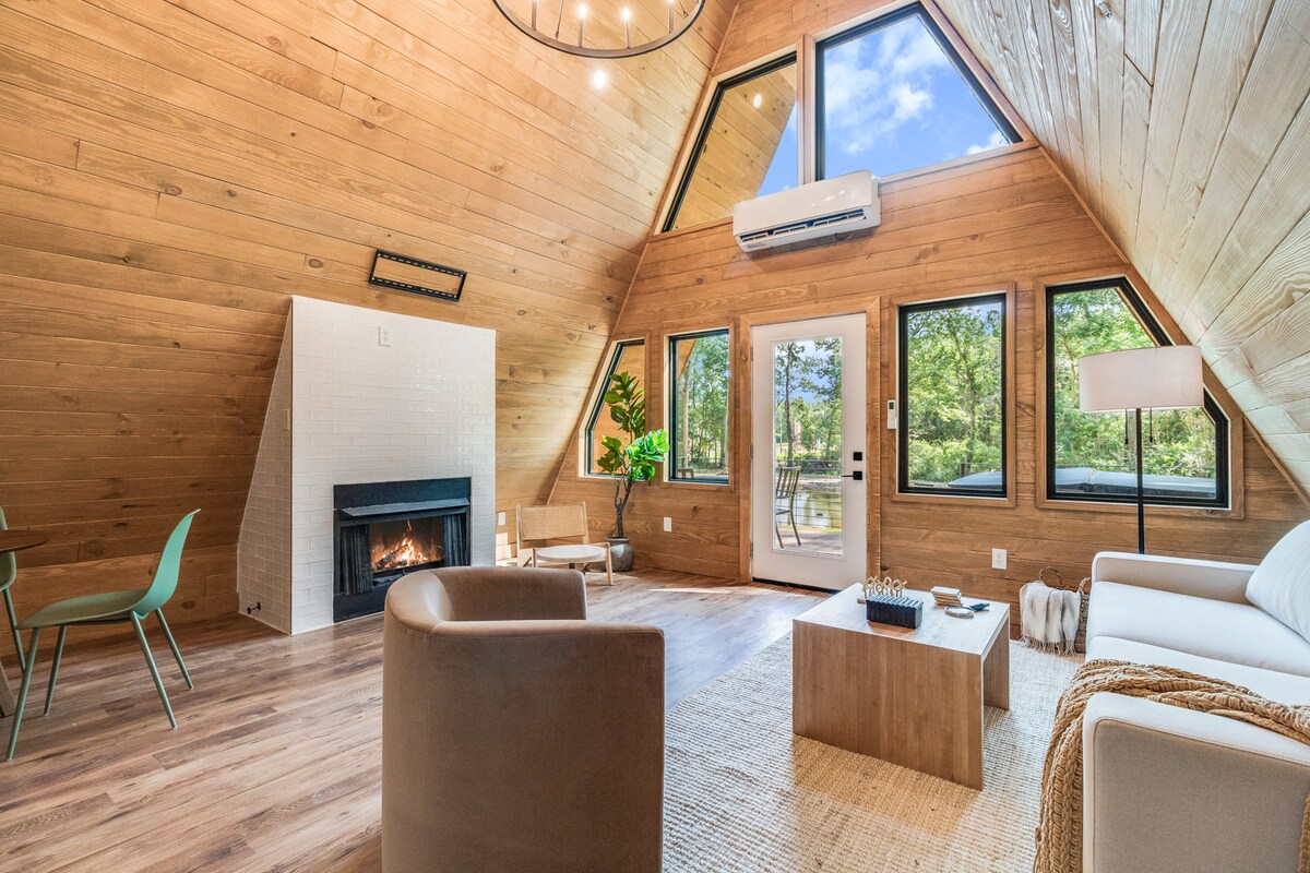 An A-frame living area is showcased with wooden walls and a sloped ceiling. Large windows invite natural light and provide views of the outdoors. A cozy seating arrangement includes a white sofa and two chairs around a wooden coffee table, with a fireplace adding warmth to the space.