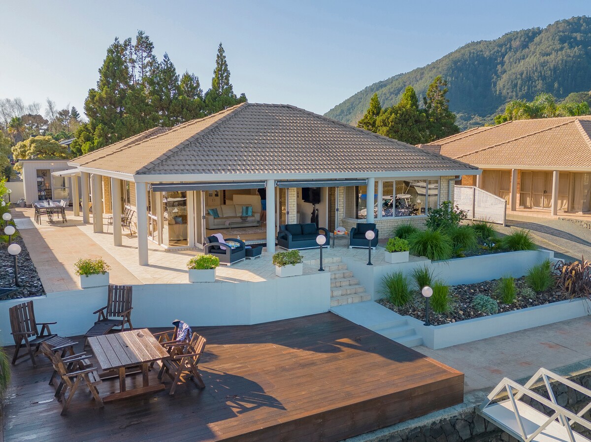 The exterior of a spacious executive home is presented, showcasing a covered patio area with comfortable seating and a dining table. Lush greenery and well-maintained landscaping frame the property, while a wooden deck extends toward the canal's edge, inviting relaxation and enjoyment of the views.
