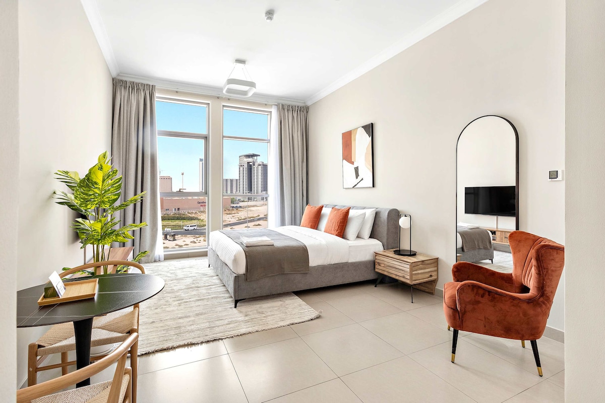 A modern studio space is presented with a spacious double bed, accompanied by a comfortable orange armchair. A small round table is positioned next to the bed, while large windows allow natural light to illuminate the room, offering views of the city beyond.