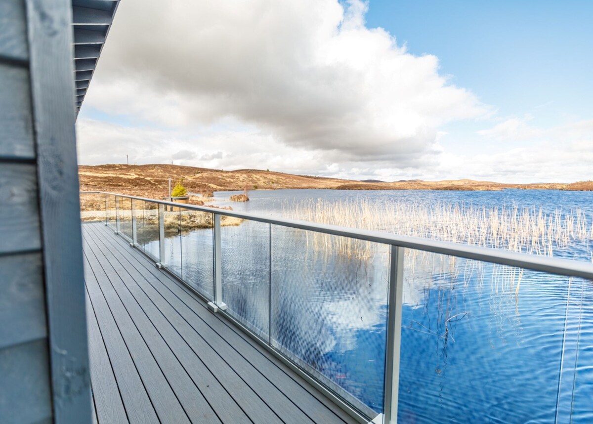 A spacious deck, presented in a modern grey design, overlooks the calm waters of Loch na Beinne Moire. The surface is adorned with wooden boards, and a glass railing provides an unobstructed view of the serene landscape, characterized by gentle hills and expansive blue skies.