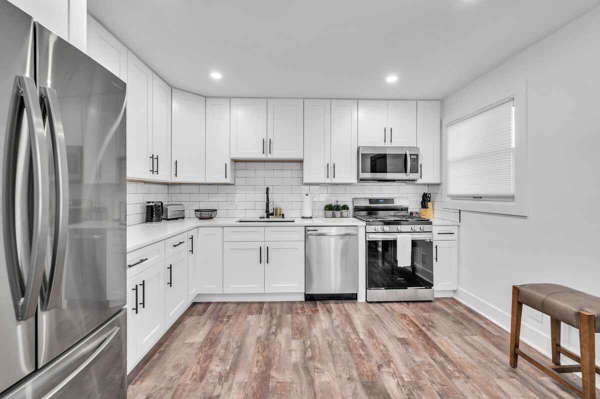 The kitchen features modern white cabinetry with stainless steel appliances, including a refrigerator and oven. A clean tile backsplash complements the spacious layout. Natural light enters through a window adorned with a simple covering, illuminating the wooden floor and a stool positioned against the cabinetry.