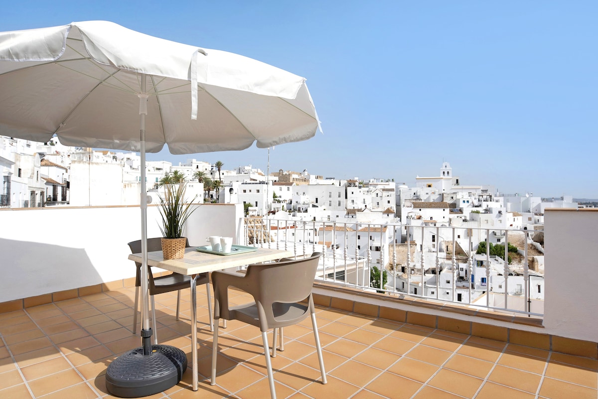 A private open terrace features a dining table surrounded by four chairs, shaded by a large umbrella. The view showcases the white buildings of Vejer against a clear blue sky, creating a serene and spacious outdoor area.