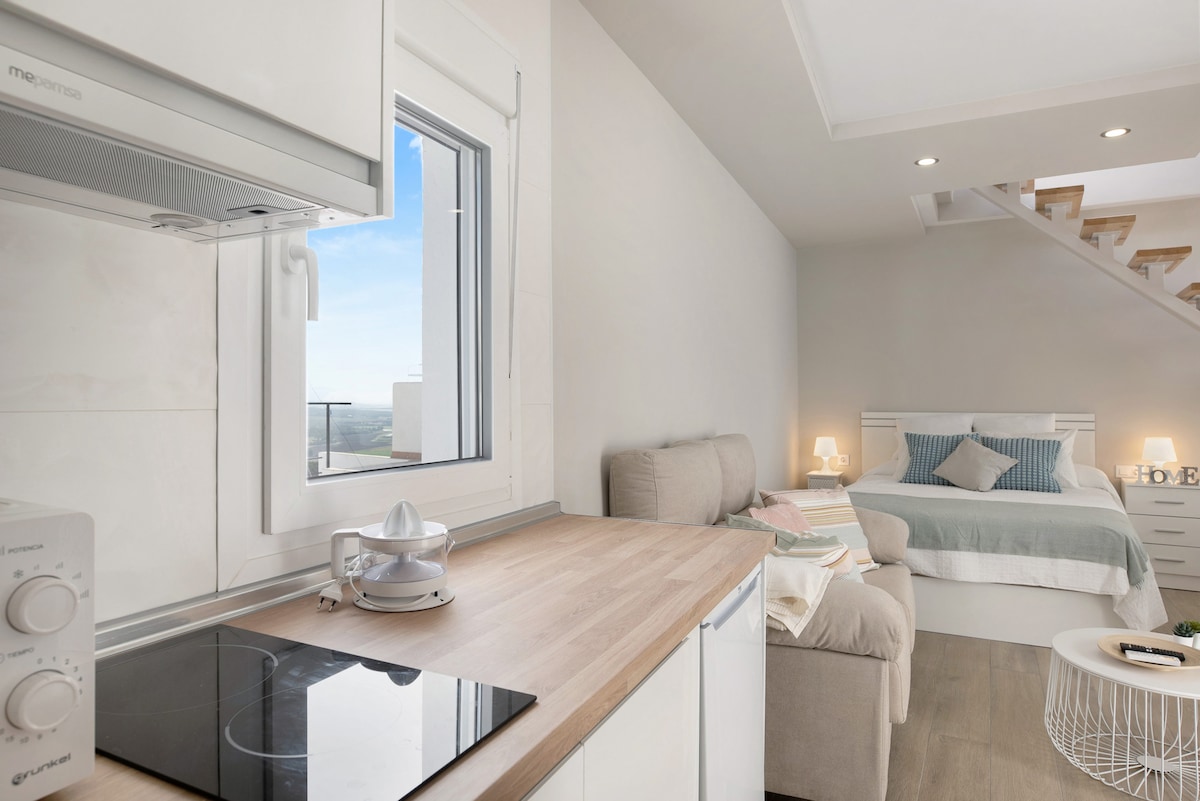 A well-arranged kitchen area features a compact stovetop and a modern microwave, with natural light coming through a nearby window. The open living space includes a spacious bed and a cozy seating area, creating a functional layout for relaxation.