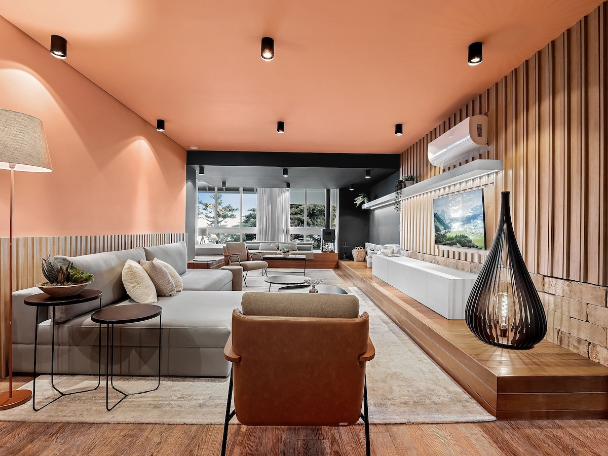 The living area features a modern layout with a large sofa and a pair of side tables. A floor lamp provides soft lighting, while a wooden accent wall complements the unique ceiling color. A large TV sits mounted on the wall, surrounded by ample natural light from expansive windows.