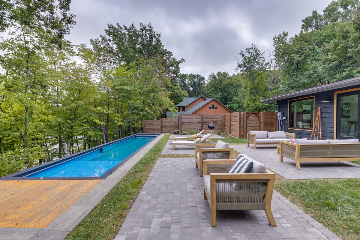 An outdoor space is showcased, featuring a seasonal pool bordered by a stone patio. Comfortable seating areas with light-colored cushions are positioned nearby, surrounded by greenery. A fenced area offers privacy, and trees provide a natural backdrop to the relaxing environment.