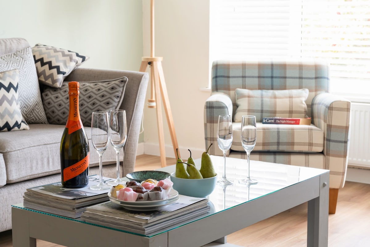 A glass coffee table is set with a plate of assorted sweets and fresh pears. Nearby, a bottle of sparkling wine is accompanied by four champagne flutes. A comfortable armchair with a plaid pattern complements a cozy gray sofa adorned with patterned cushions, while natural light enters through the window.
