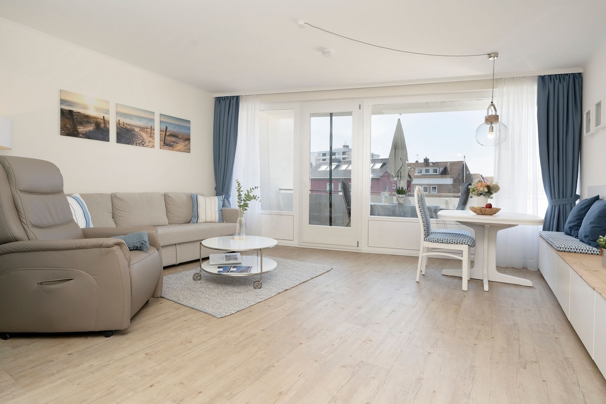 A bright living area features a comfortable leather armchair, a light-coloured sofa, and a round glass coffee table. Large windows allow natural light to fill the space, with soft curtains framing the view. The dining area includes a white table and chairs, decorated with a vase of flowers.
