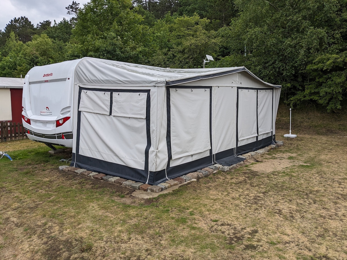 A spacious rental caravan is parked on a grassy area, surrounded by trees. An attached canopy extends from the side, featuring fabric walls for added privacy. The structure is designed for comfort, with ample space for relaxation outdoors.