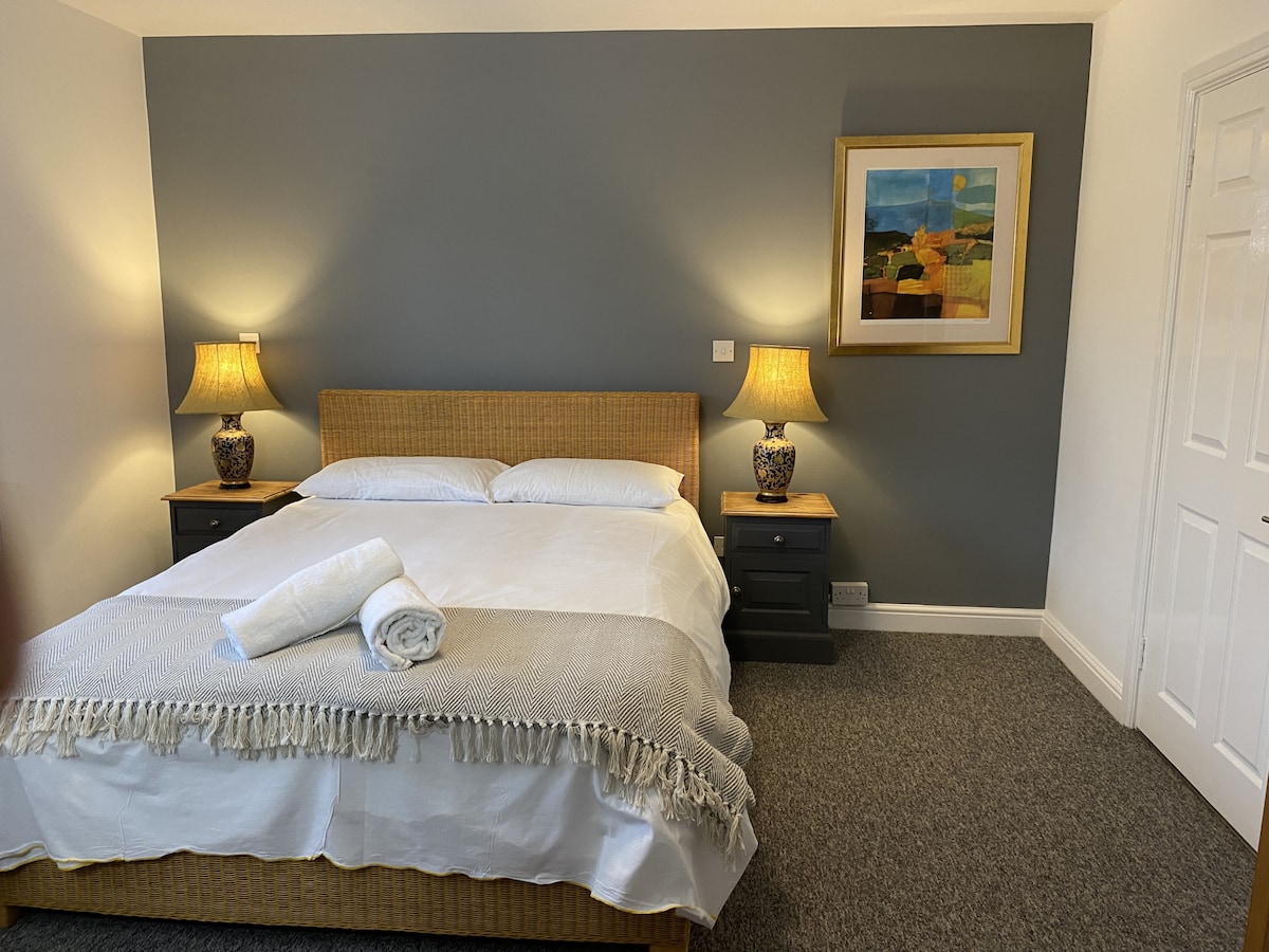 A spacious sleeping area is furnished with a large bed, complemented by two bedside tables with stylish lamps. The walls feature a soothing grey tone, and a colorful artwork hangs above the bed, adding an artistic touch to the serene atmosphere.