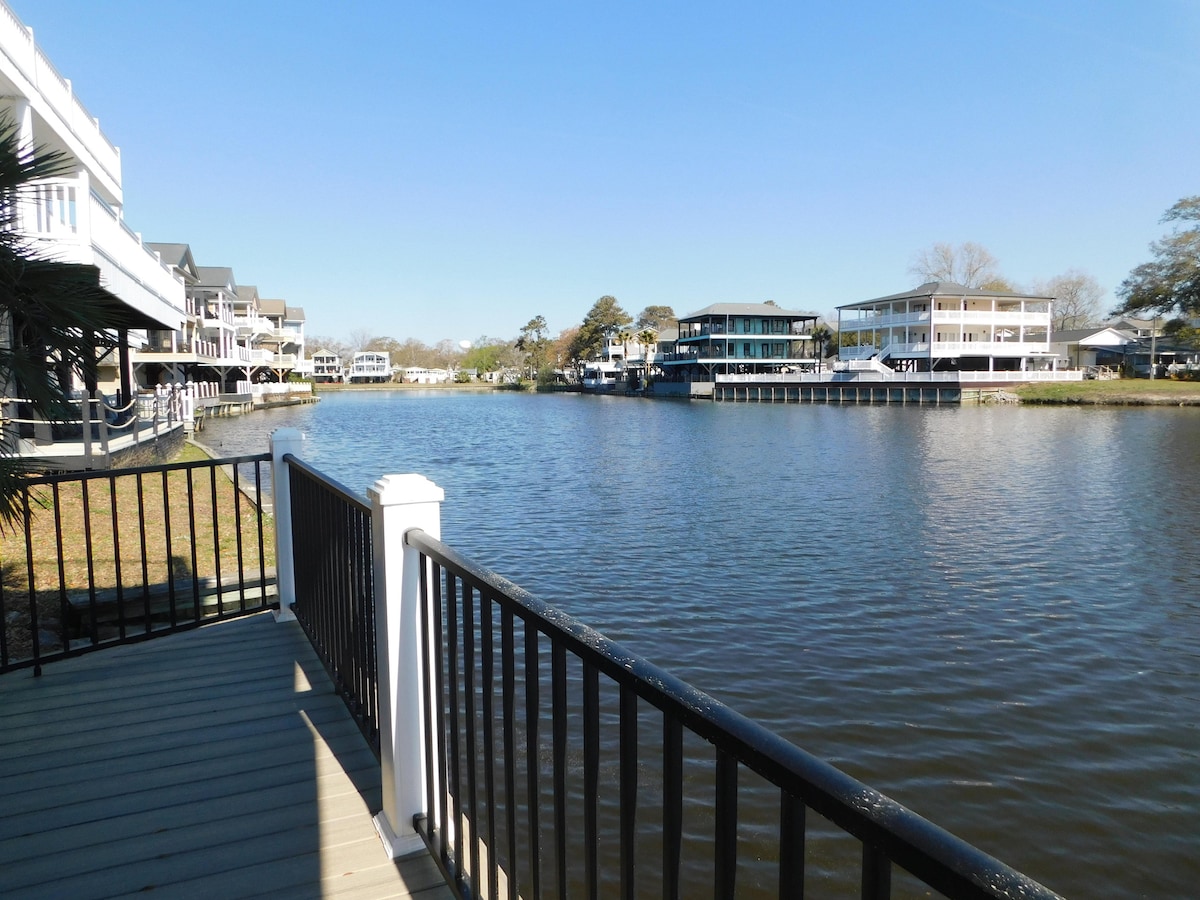 A serene water view is presented from the deck, featuring calm lake waters gently reflecting nearby houses. The deck railing enhances the perspective of the landscape, with clear blue skies above and hints of greenery along the shoreline.