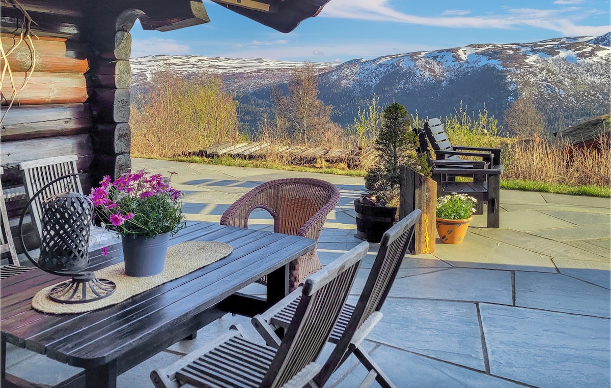 An outdoor terrace features a wooden dining table and chairs, complemented by potted flowers. The expansive view reveals rolling hills and snow-capped mountains under a clear sky, creating a serene backdrop for gathering or relaxation.
