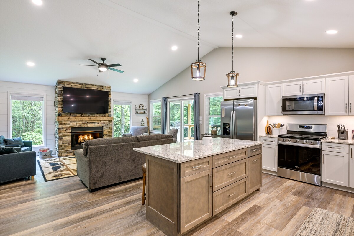An open-concept living space features a modern kitchen island with seating, flanked by stainless steel appliances. A stone fireplace serves as a focal point, with a large television mounted above. Natural light floods the room through windows overlooking the surrounding greenery.