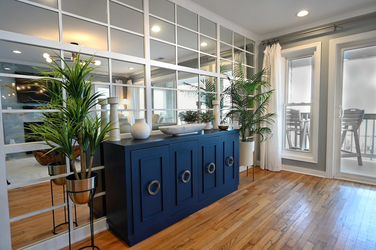 A modern entryway features a navy blue console table with circular handles, flanked by potted plants. Natural light filters through large windows, enhancing the warm wooden floors. A sliding glass door leads to a balcony, adding to the inviting atmosphere of the space.