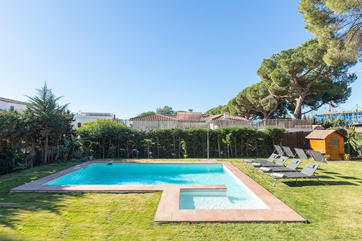 A spacious salt-water pool is set within a lush green garden, framed by tall trees. Six lounge chairs are arranged around the pool area on the well-maintained grass, alongside a small wooden shed adding a charming touch to the outdoor space.