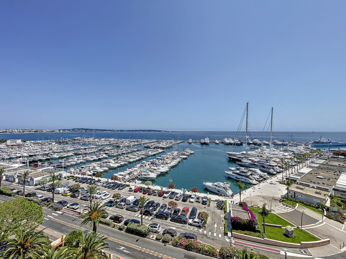 A panoramic view showcases a bustling marina filled with various yachts and boats. Rows of parked cars are visible along the waterfront, complemented by lush greenery and vibrant flowers lining the edges. The serene blue waters extend towards the horizon under a clear sky.
