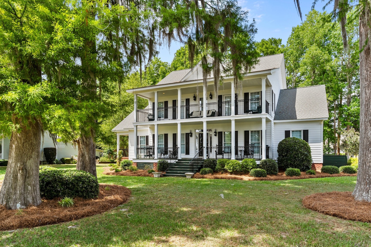 The custom colonial-style home is framed by lush greenery, showcasing a welcoming facade with multiple stacked porches. The well-maintained landscaping features manicured bushes and large trees, creating a serene outdoor environment. The upper and lower porches include black railings, enhancing the home's charm.