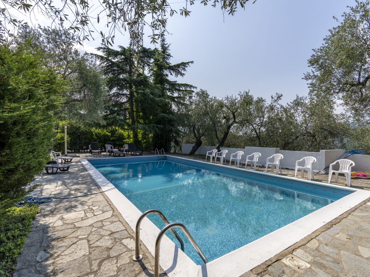 A fenced swimming pool, measuring 10 by 5 meters, is set in a natural garden surrounded by olive trees. The pool is equipped with a set of lounge chairs and sun loungers, providing a relaxing outdoor space. The area is easily accessible from the residence.