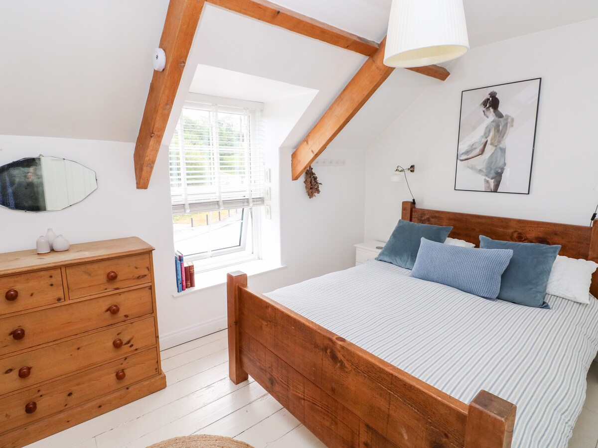 A bright bedroom is presented with exposed wooden beams and a double bed featuring a wooden frame. Pillows in soft blue tones are arranged on the bed. A window with interior shutters allows natural light, while a wooden dresser and a framed artwork add subtle decor.