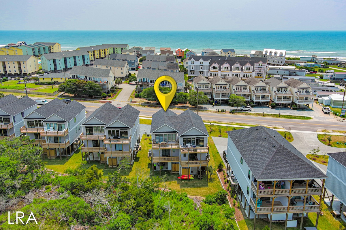 An aerial view captures the property nestled within a coastal community. The ocean is visible in the background, with nearby buildings and walkways leading to the beach. Lush green grass surrounds the house, which features a dark roof and multiple decks, providing a sense of space and accessibility.