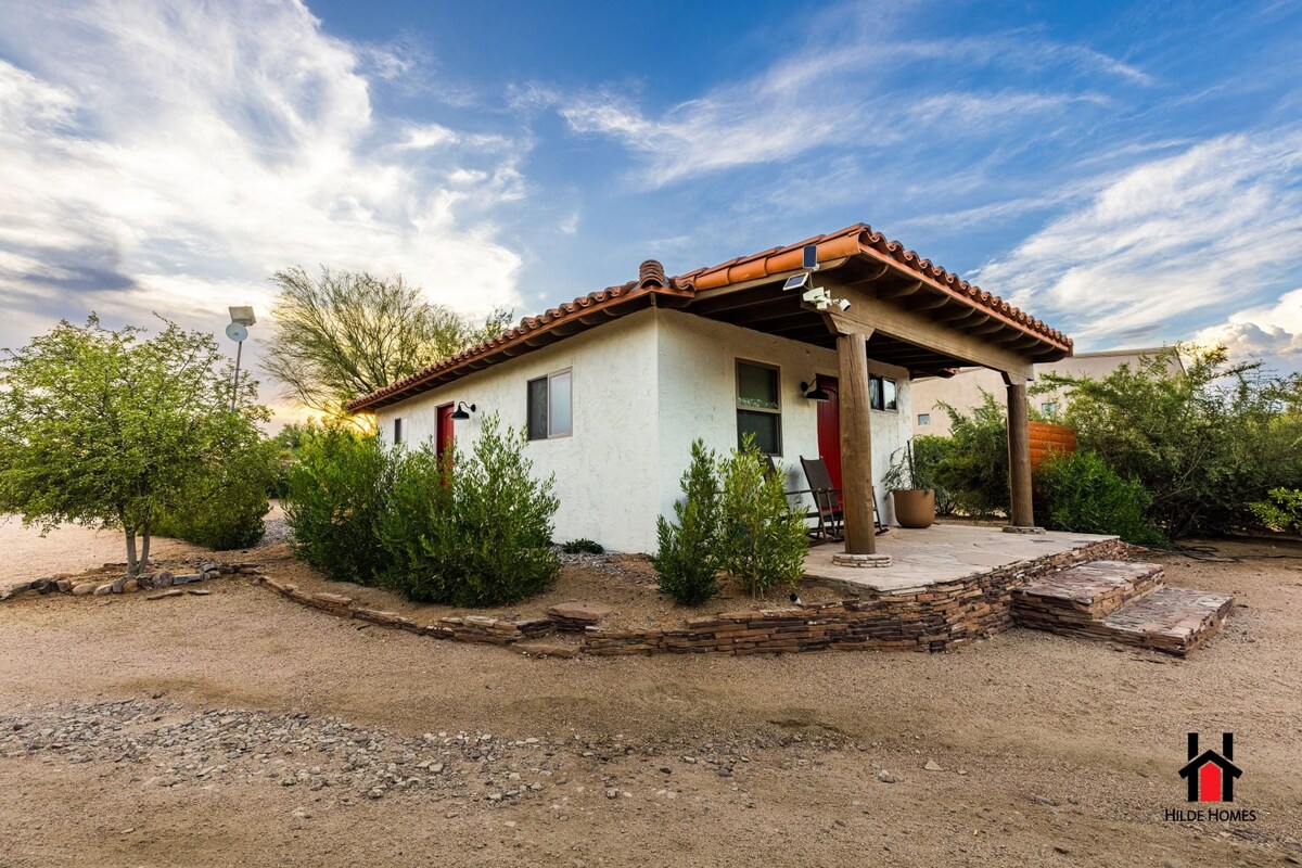 The exterior of the Cardinal Casita is highlighted by a charming, single-story structure surrounded by native desert vegetation. The entryway features a spacious patio area with stone pathways and a couple of lounge chairs, inviting guests to relax while enjoying the serene desert landscape.