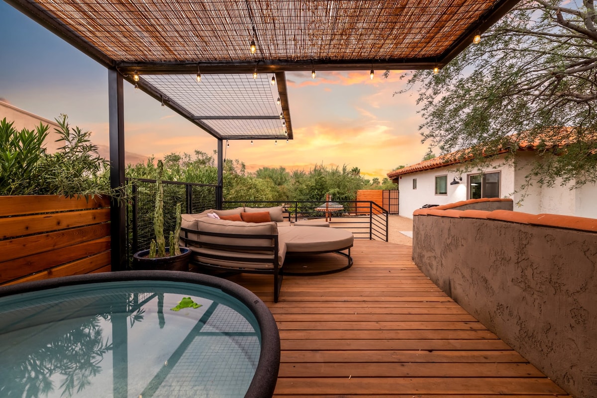 A spacious deck highlighted by a chic sofa lounge is adorned with market lights, providing a cozy ambiance for relaxation. A cowboy tub is positioned nearby, surrounded by lush greenery, offering a refreshing escape while overlooking the desert landscape at sunset.