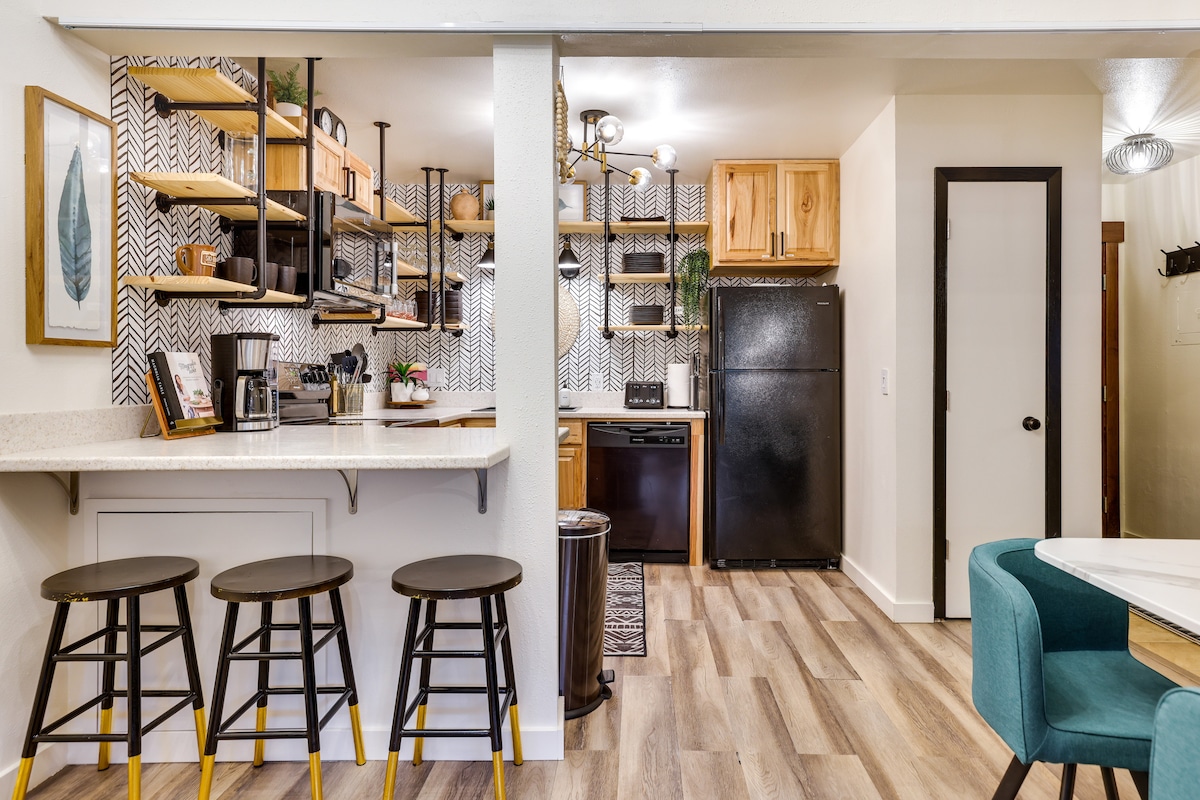The kitchen area features open shelving with decorative items and cooking essentials. A countertop with three stools provides seating, while a refrigerator and dishwasher are visible in the background. The modern design is complemented by stylish wall patterns and warm wood cabinetry.