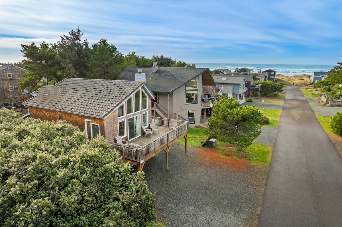 An aerial view captures a contemporary beach house with a large deck, positioned amongst lush greenery. The ocean is visible in the background, while nearby homes are seen along a quiet road, emphasizing the tranquil coastal setting.