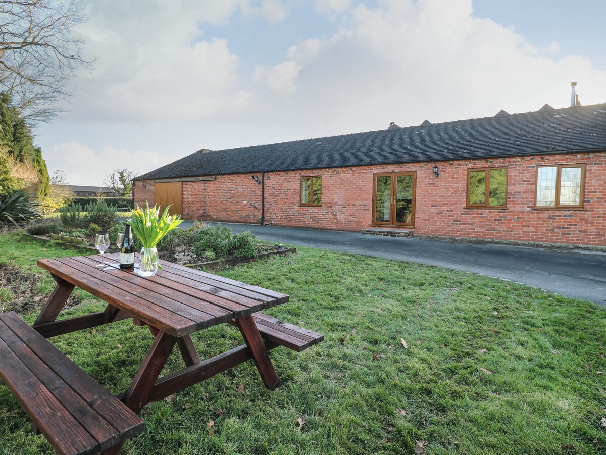 A wooden picnic table sits on a lush green lawn, accompanied by a bouquet of flowers and two glasses. The cottage's brick facade is visible in the background, featuring multiple windows that allow natural light to fill the interior.